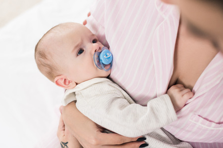 partial view of little baby with pacifier on mothers hands