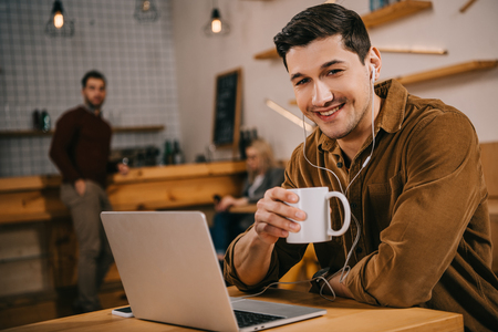handsome man in earphones holding cup of coffee near laptop in cafeの写真素材