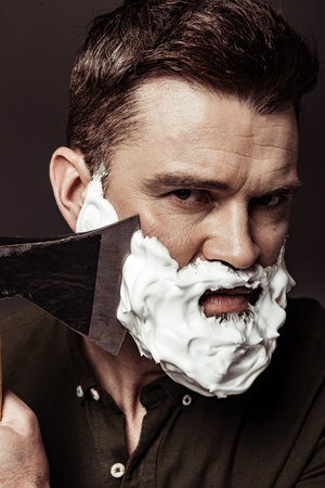 close up of serious handsome bearded man shaving with ax while looking at camera isolated on brownの写真素材