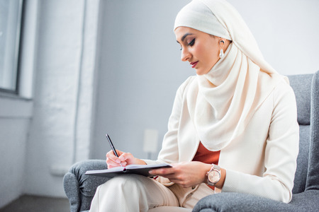beautiful young muslim woman sitting in chair and writing in notebookの写真素材