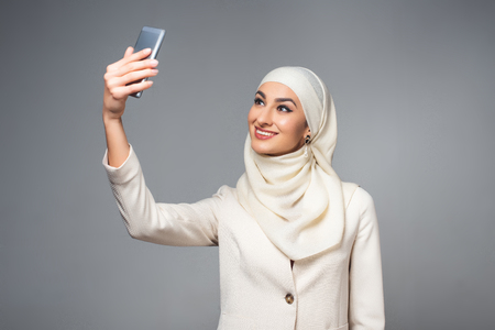 beautiful smiling muslim woman taking selfie with smartphone isolated on greyの写真素材