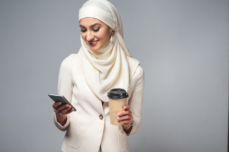 smiling young muslim woman holding coffee to go and using smartphone isolated on greyの写真素材