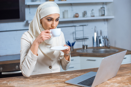 young muslim woman drinking coffee and using laptop at homeの写真素材