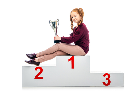 happy schoolgirl sitting on winner podium, holding trophy cup and looking at camera isolated on whiteの写真素材
