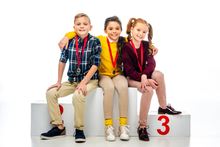 smiling kids with medals sitting on winner podium and looking at camera isolated on whiteの写真素材