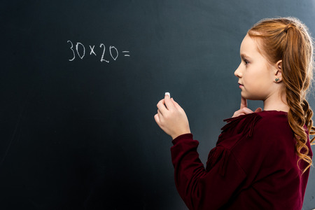 thoughtful schoolgirl holding chalk and looking at blackboard with math exampleの写真素材