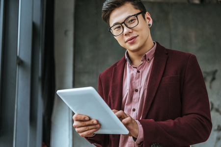 handsome kazakh businessman in eyeglasses using digital tablet and looking at camera in officeの写真素材