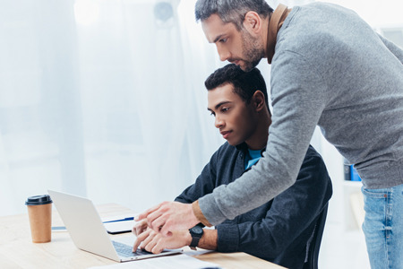 bearded mentor helping young colleague working with laptop in officeの写真素材