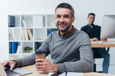 handsome smiling businessman holding coffee to go and using laptop in officeの写真素材