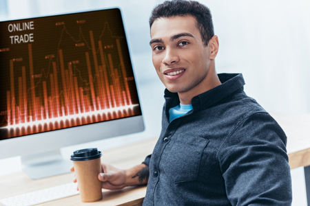 young businessman with coffee to go working with desktop computer with online trade graphs and smiling at cameraの写真素材