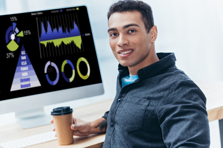 young businessman with coffee to go working with desktop computer and smiling at cameraの写真素材
