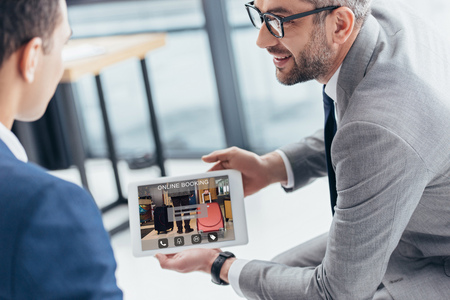 cropped shot of businessman in eyeglasses showing digital tablet with online booking app on screen to male colleague in officeの写真素材