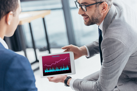 cropped shot of businessman in eyeglasses showing digital tablet with trading graphs on screen to male colleague in officeの写真素材