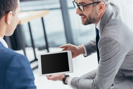 cropped shot of businessman in eyeglasses showing digital tablet with blank screen to male colleague in officeの写真素材