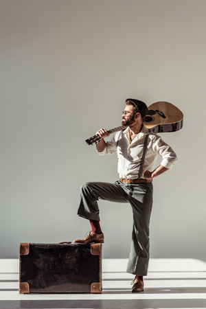 bearded man in cap putting foot on vintage suitcase and holding acoustic guitar on shoulder on grey backgroundの写真素材