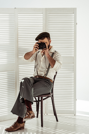 bearded man sitting on chair and taking pictures with vintage film cameraの写真素材