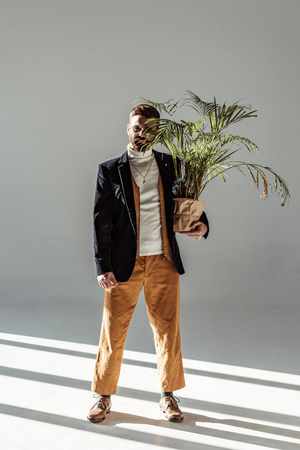 handsome bearded man in glasses holding green plant in pot and looking at camera on grey backgroundの写真素材