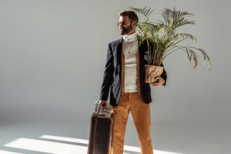 handsome bearded man in glasses holding green plant in pot and vintage suitcase on grey backgroundの写真素材