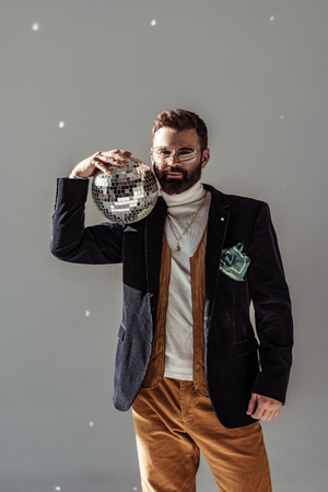 handsome bearded man in glasses holding disco ball on shoulder and looking at camera on grey backgroundの写真素材