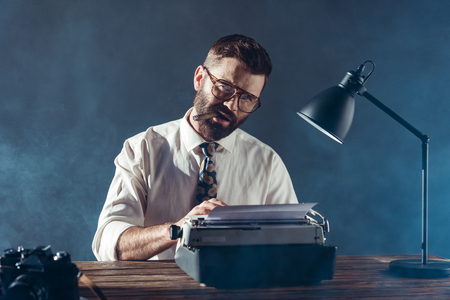 bearded journalist sitting at table, typing on typewriter, smoking and looking at camera on grey backgroundの写真素材