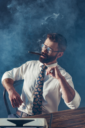 thoughtful bearded journalist in glasses sitting at table with vintage typewriter and smoking on grey backgroundの写真素材