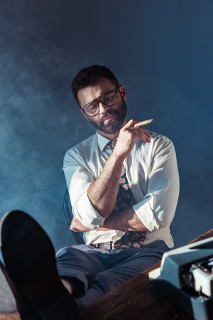 bearded journalist with closed eyes putting feet up on table and smoking on grey backgroundの写真素材