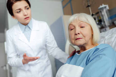 upset female doctor consoling sad senior woman lying in hospital bedの写真素材