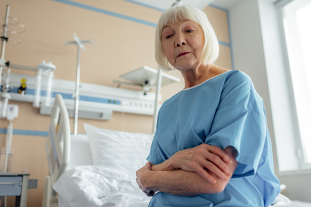 selective focus of sad senior woman with arms crossed sitting on bed in hospital wardの写真素材