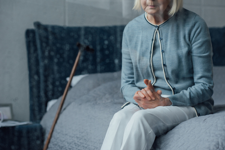 partial view of senior woman sitting on bed with folded hands at homeの写真素材