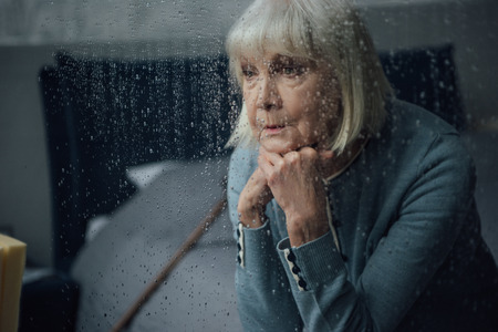 upset senior woman sitting and propping chin with hands at home through window with raindropsの写真素材