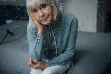 lonely senior woman sitting and looking at camera through window with raindropsの写真素材