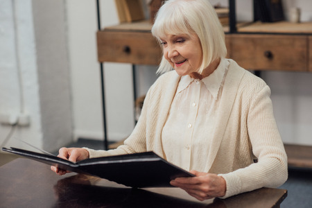 smiling senior woman sitting at table and looking at photo album at homeの写真素材