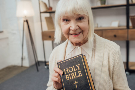 smiling senior woman looking at camera and holding holy bible at homeの写真素材