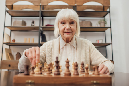 senior woman sitting at table, looking at camera and playing chess at homeの写真素材