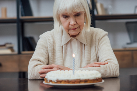 upset senior woman sitting at table and looking at birthday cake with burning candle at homeの写真素材