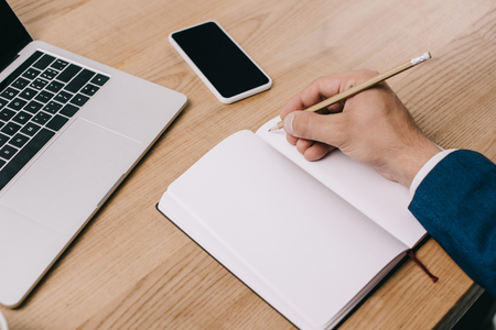 cropped view of businessman writing in notebook at workplace with gadgetsの写真素材