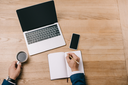 cropped view of businessman writing in notebook at workplace with coffee and digital devicesの写真素材
