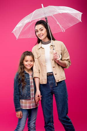mother with umbrella and adorable happy daughter holding hands and smiling at camera isolated on pinkの写真素材
