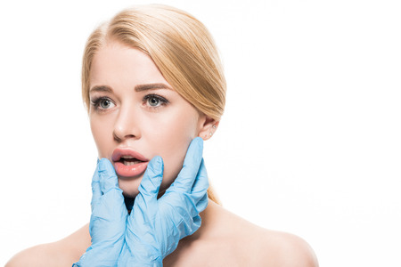 cropped shot of cosmetologist in latex gloves touching face and lips of young woman isolated on...
