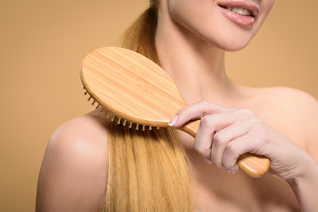 cropped shot of naked blonde girl brushing hair with wooden hairbrush isolated on beigeの写真素材