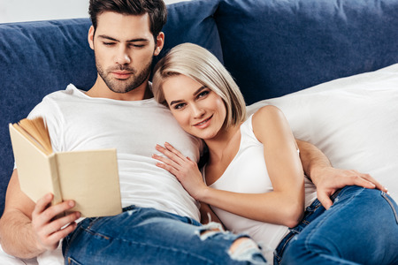 selective focus of attractive girlfriend and handsome boyfriend laying while smiling and reading bookの写真素材