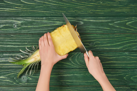 cropped view of woman cutting pineapple with knife on green wooden tableの写真素材