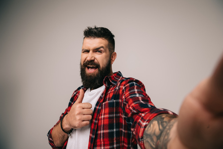 excited man in checkered shirt showing thumb up while taking selfie, isolated on greyの写真素材