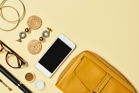Top view of earrings, glasses, eyeshadow, smartphone, mascara, bracelets, cosmetic brush and bag on...