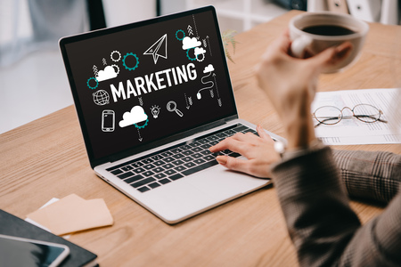 cropped view of businesswoman with coffee cup typing on laptop with marketing iconsの写真素材