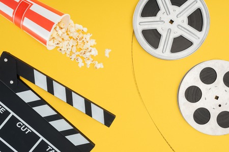 top view of clapperboard, overturned striped bucket with popcorn and film reels with cinema tape isolated on yellowの写真素材