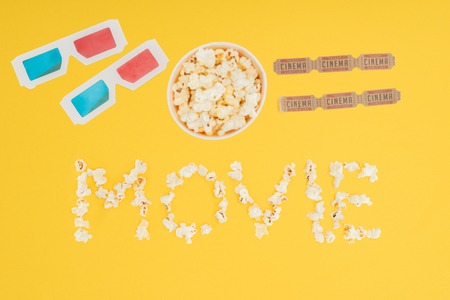 top view of 3d glasses, bucket with popcorn, cinema tickets and "movie" lettering made with popcorn isolated on yellowの写真素材