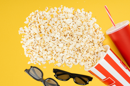 top view of 3d glasses, red disposable cup with straw and overturned striped bucket with popcorn isolated on yellowの写真素材