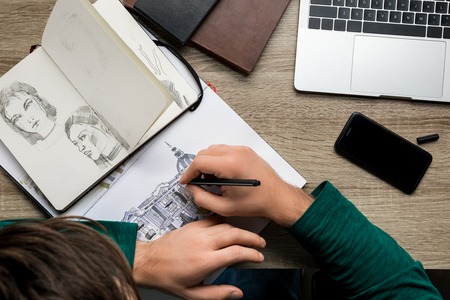 overhead back view of man drawing in album on wooden table next to laptop and smartphoneの写真素材