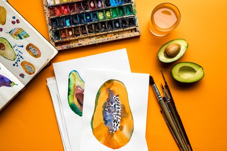 top view of colored paints, paintbrushes, sketchbook, drawings with avocado and papaya on yellow...